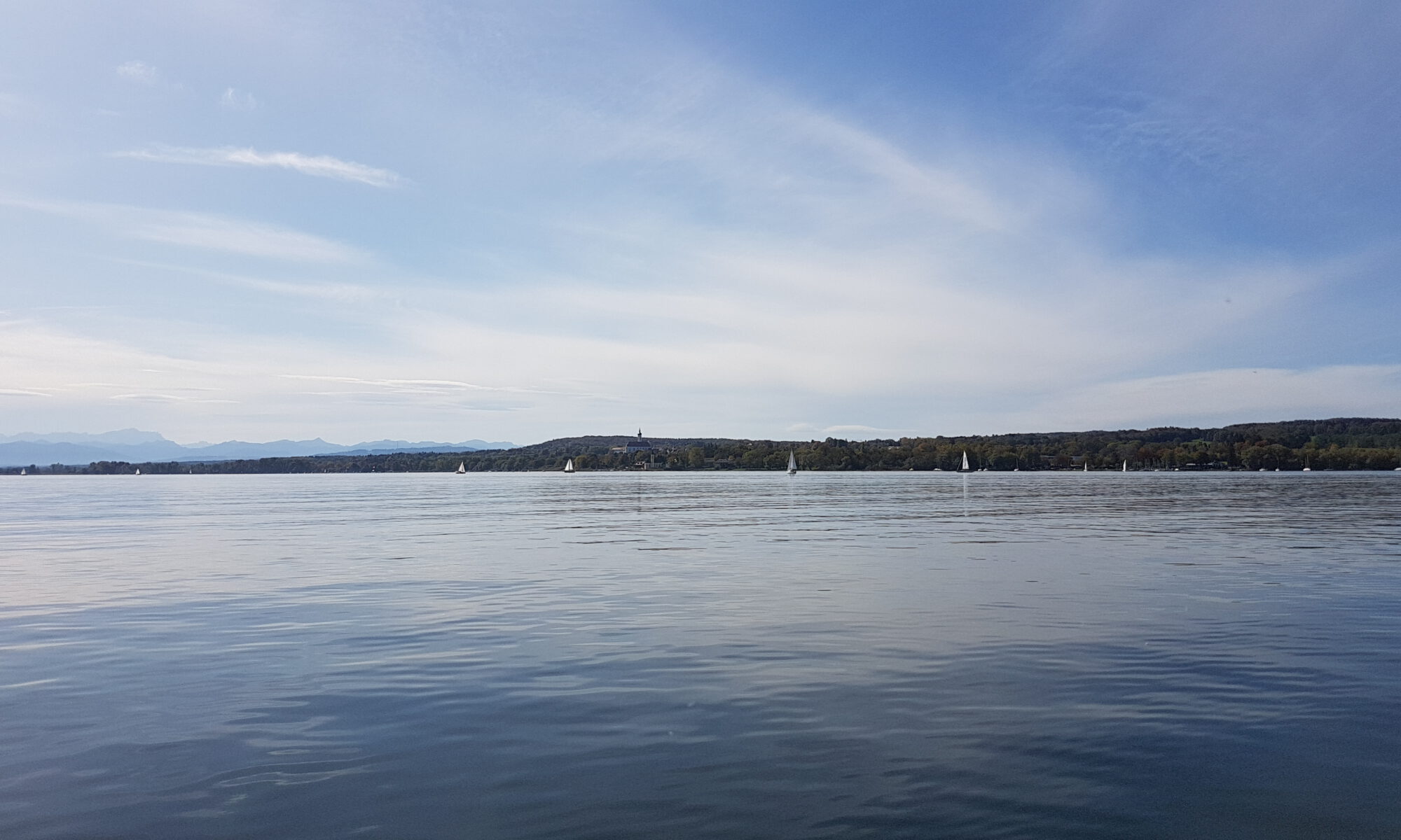 Ruhiger See mit einigen Segelbooten, bewaldetem Ufer und Bergen am Horizont unter leicht bewölktem Himmel