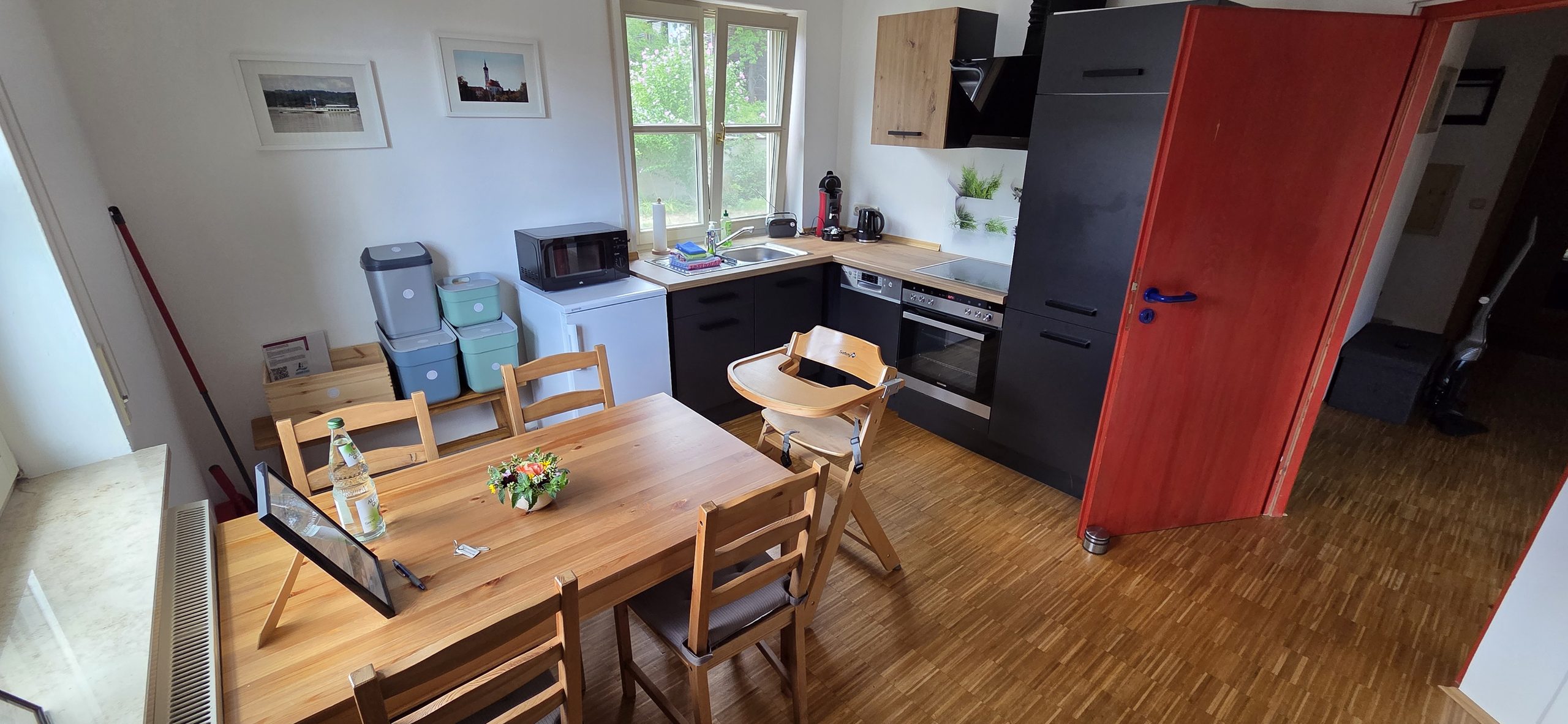 Offene Küche mit Esstisch aus Holz, dunkler Küchenzeile, Kinderhochstuhl und auffälliger roter Tür