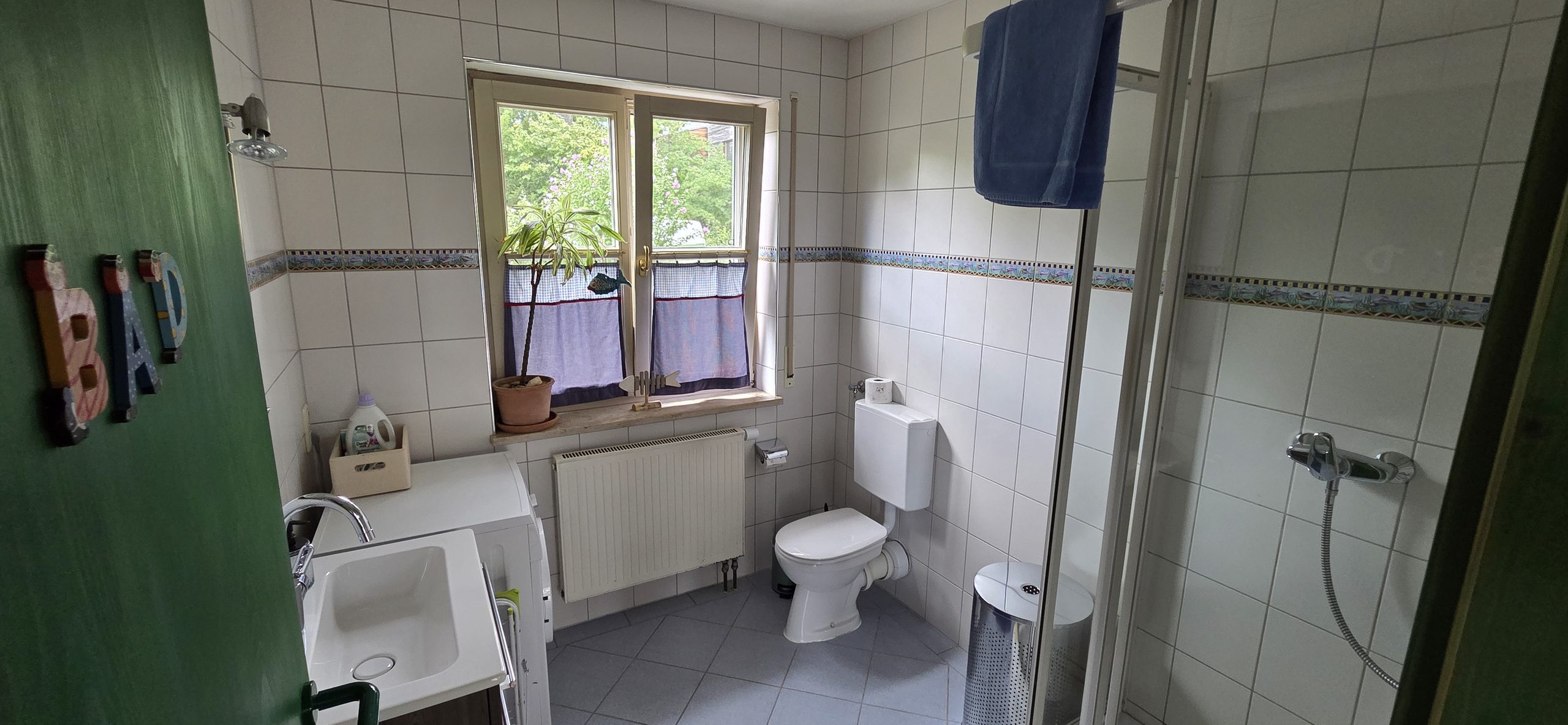 Helles, weiß gefliestes Badezimmer mit Waschbecken, WC, Dusche und einem Fenster